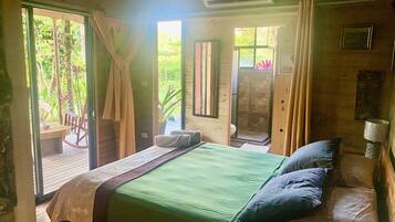 Quarto com cama, uma mesinha e uma cadeira. Há uma janela com cortinas, uma porta e um aparelho de ar condicionado.