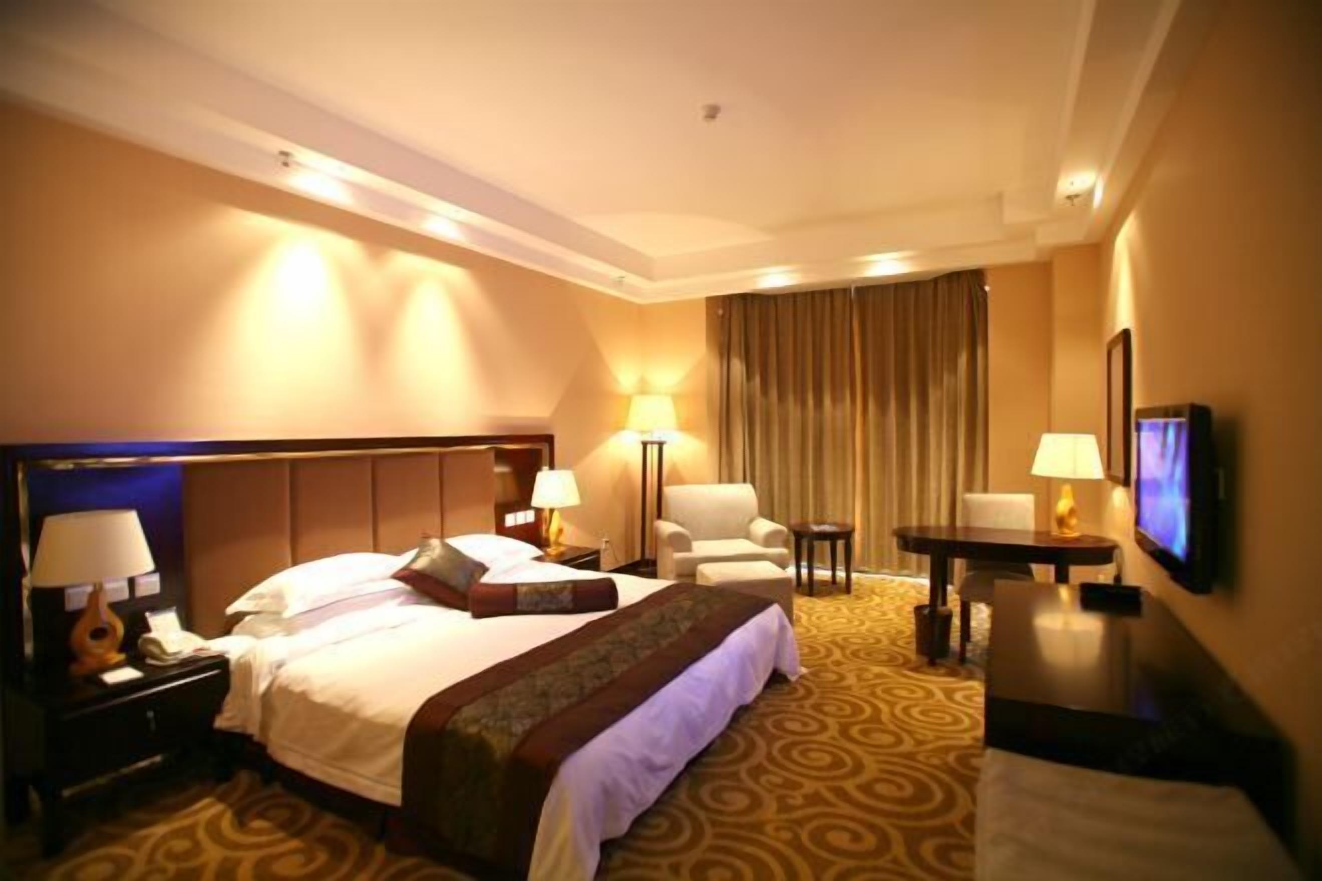 Ein Hotelzimmer mit einem großen Bett, zwei Nachttischen mit Lampen, einem Sessel, einem Schreibtisch mit Fernseher und einem Teppichboden.