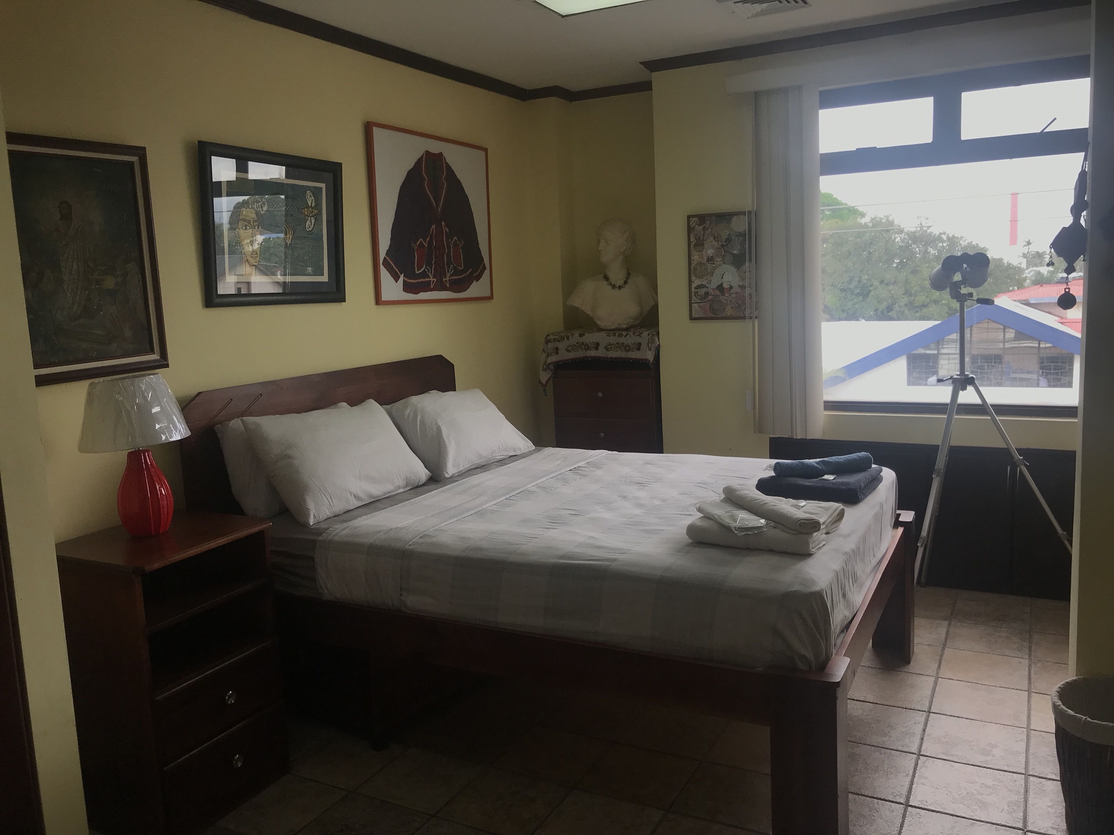 A bedroom with a bed, a nightstand, a lamp, a window with a view, and a tripod.