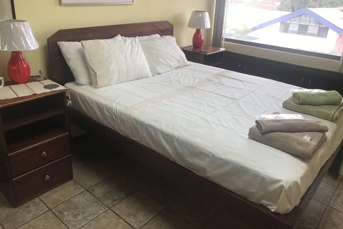 A neatly made bed with white linens, a wooden nightstand with a red lamp, and a window with a view of buildings outside.