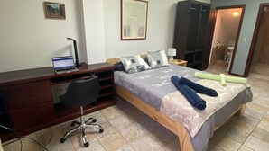 A bedroom with a bed, a desk with a laptop, a chair, a wardrobe, and a bathroom visible through an open door.