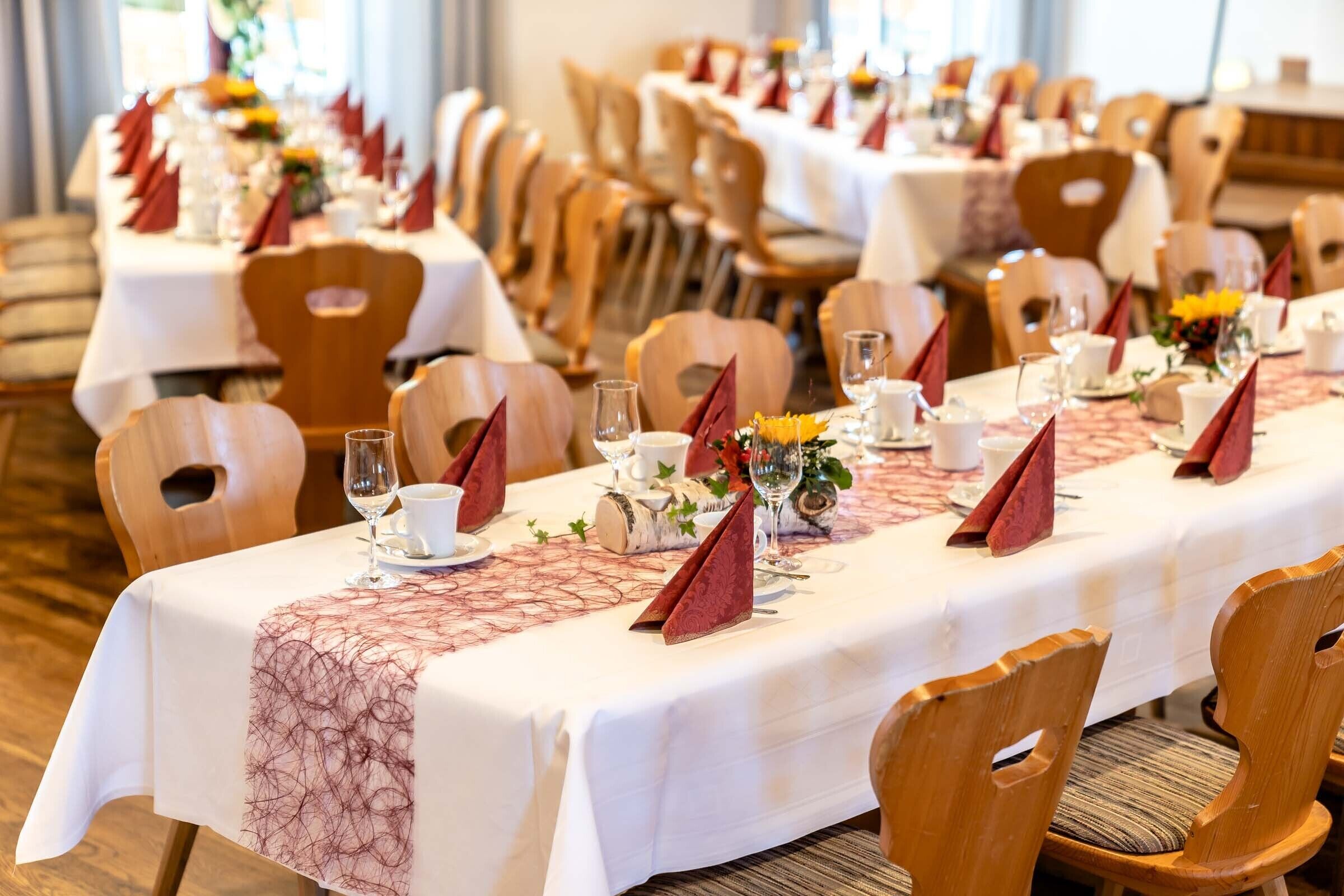 A banquet hall with long tables set for a formal event, featuring white tablecloths, wooden chairs, and red napkins.