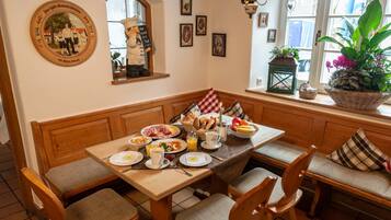 Une table en bois, des chaises et un banc sont disposés pour le petit-déjeuner. On y trouve des assiettes remplies de nourriture, des tasses et un panier de pain.