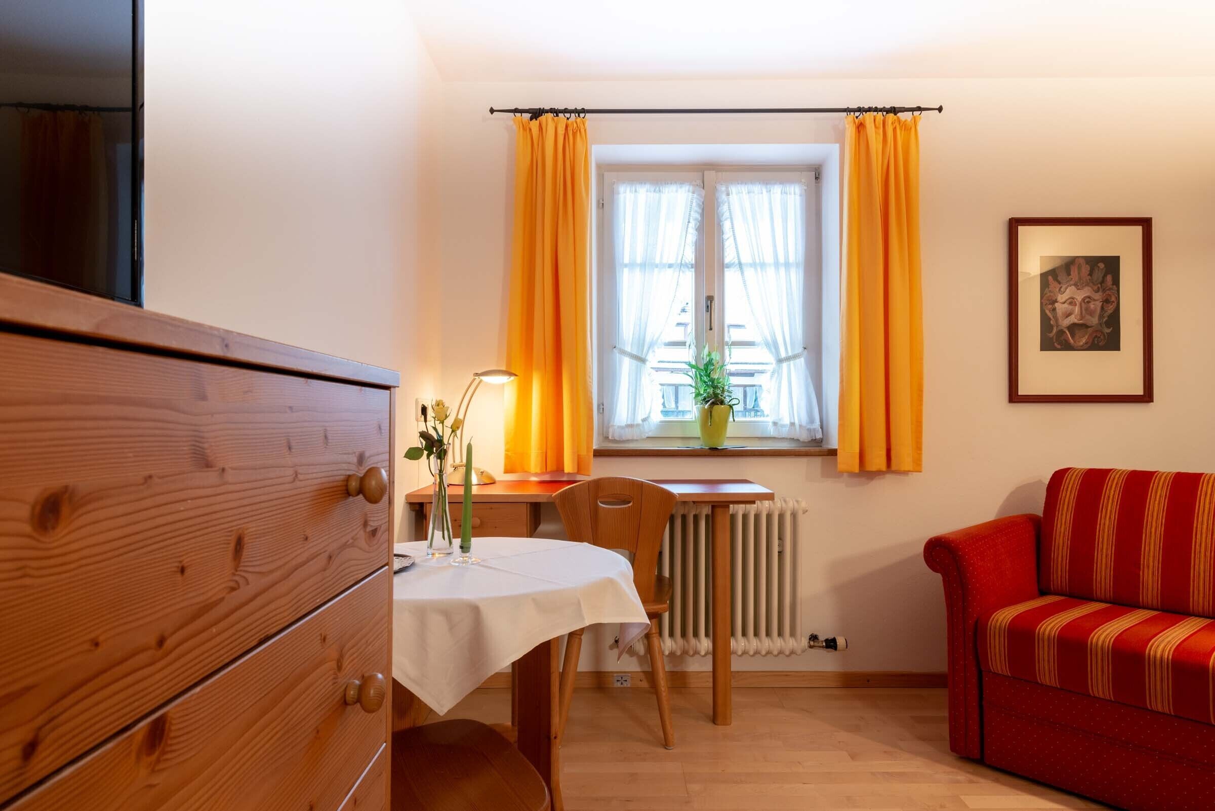 A room with a wooden dresser, a table with a white tablecloth, a red sofa, and a window with yellow curtains.