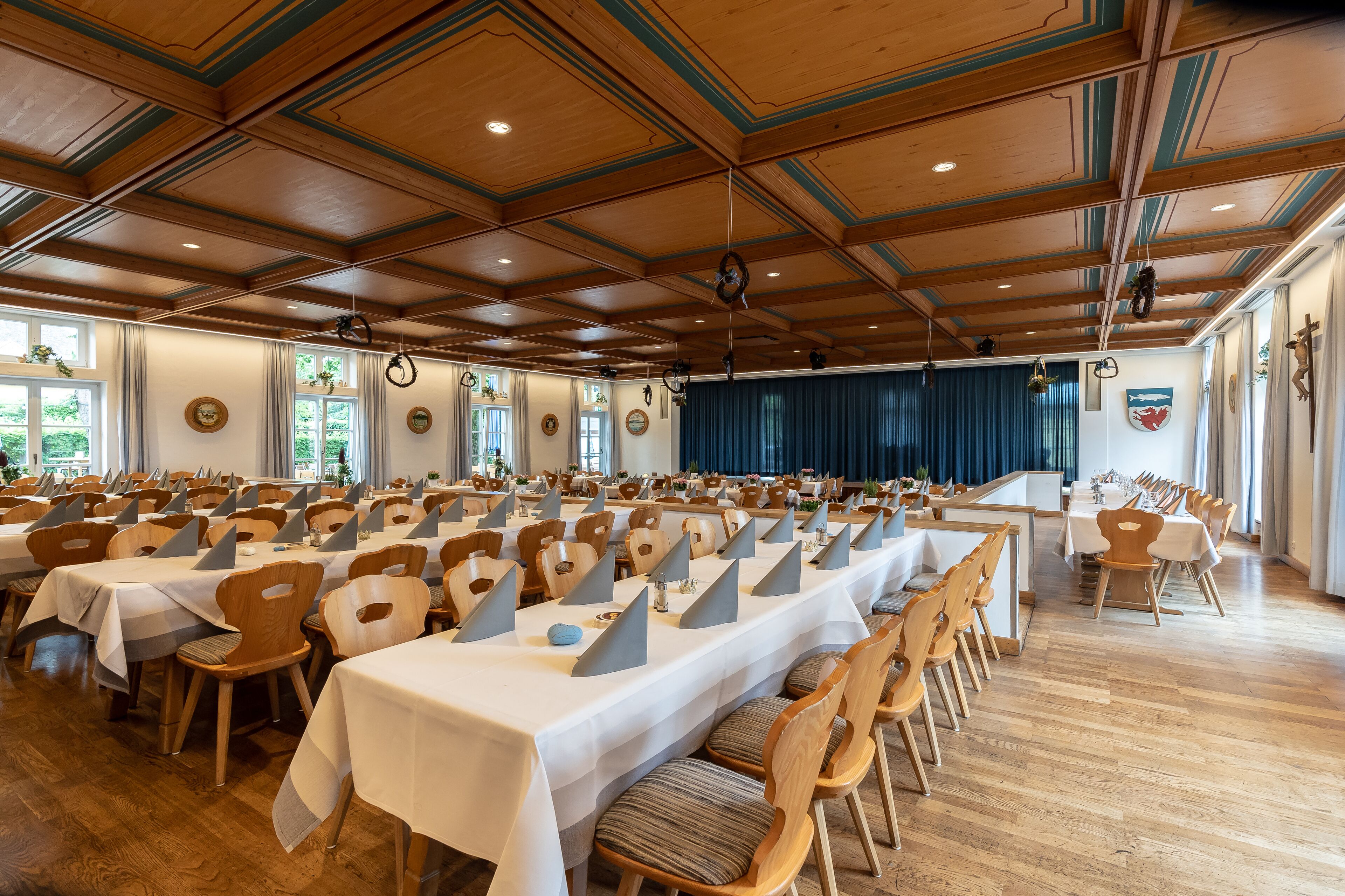Een ruime balzaal met lange tafels gedekt voor een diner, houten stoelen en een hoog plafond met houten balken.