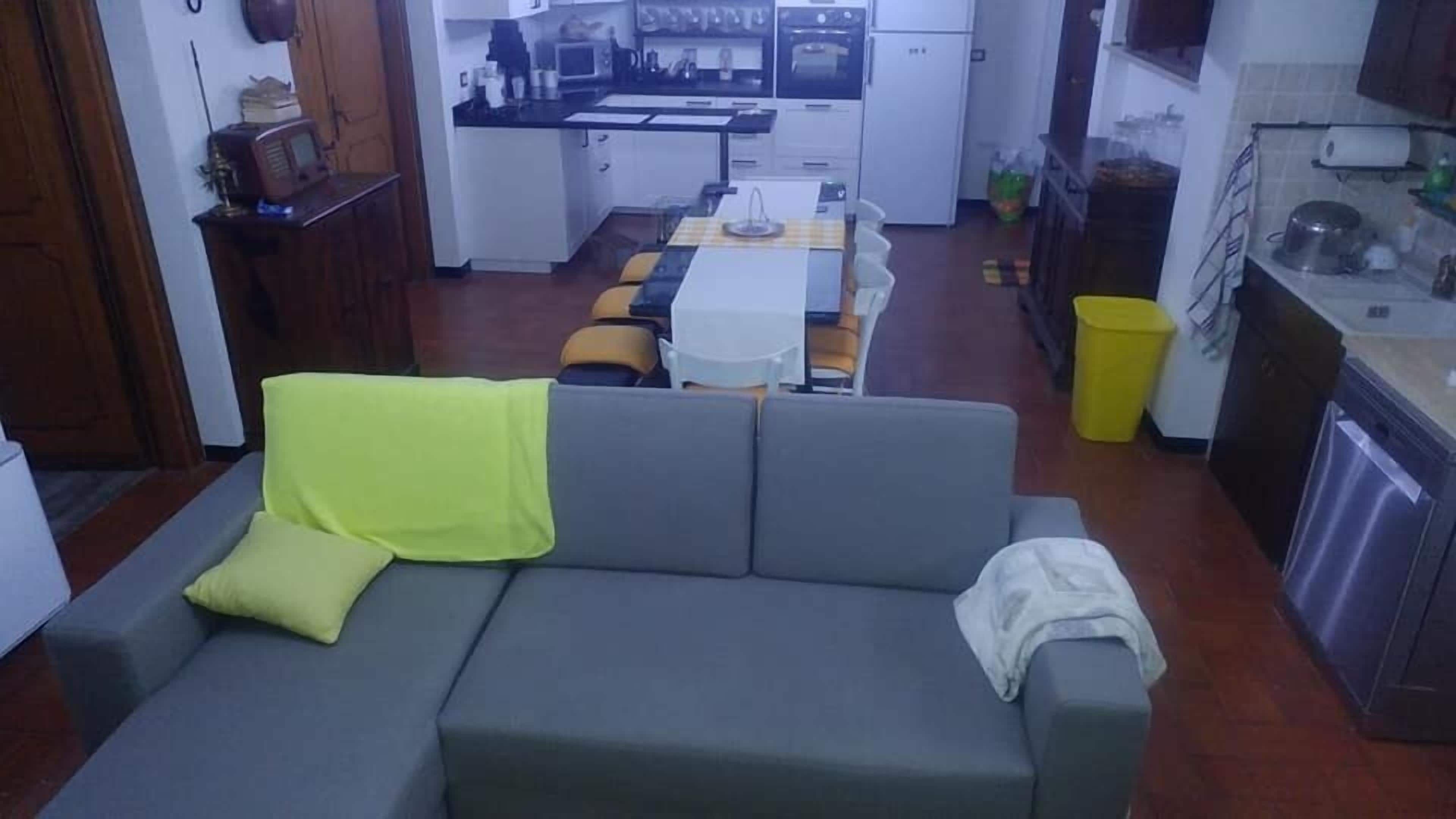Una sala de estar con un sofá gris, una manta amarilla y una manta blanca. En el fondo, se ve una cocina con una mesa y sillas.