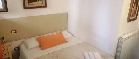 Un lit bien fait, avec un oreiller orange et une serviette blanche pliée, un tableau encadré au mur et un radiateur également fixé au mur.