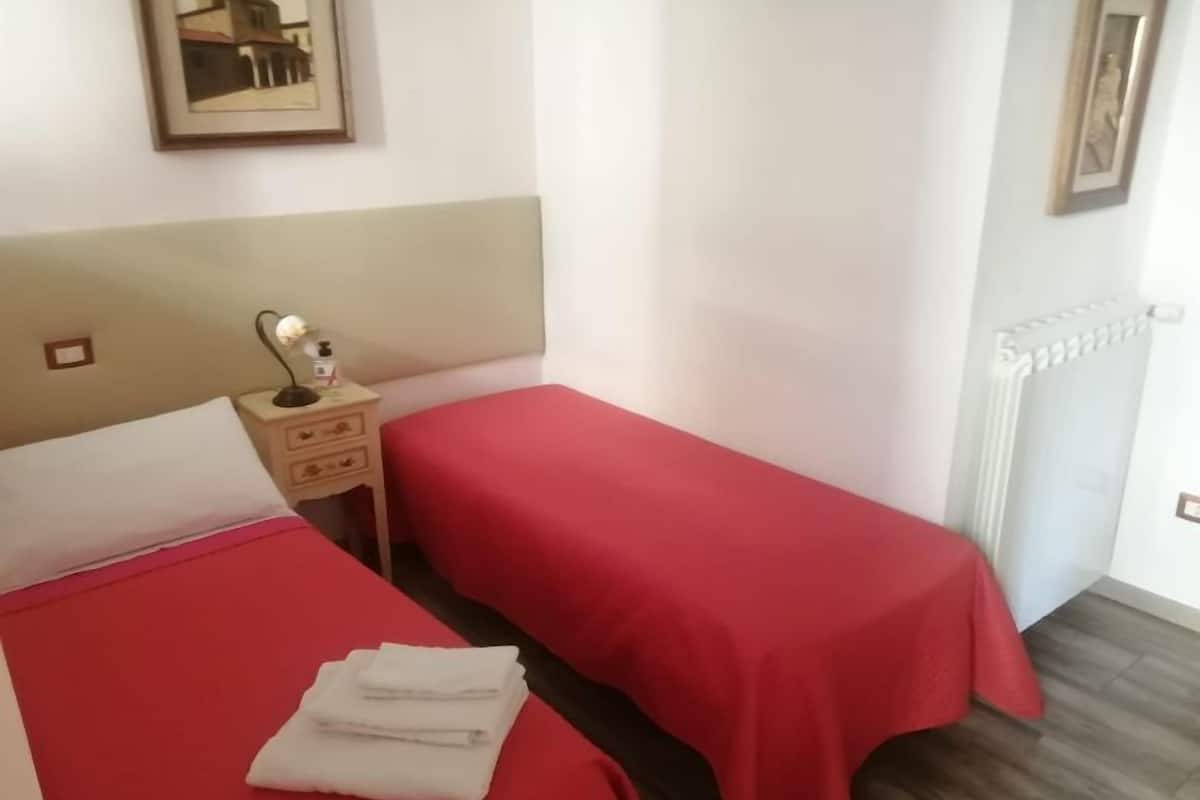 A room with two beds, each with a red bedspread and white pillows. There is a nightstand with a lamp and a small table with a bottle and a box. A framed picture hangs on the wall above the beds.
