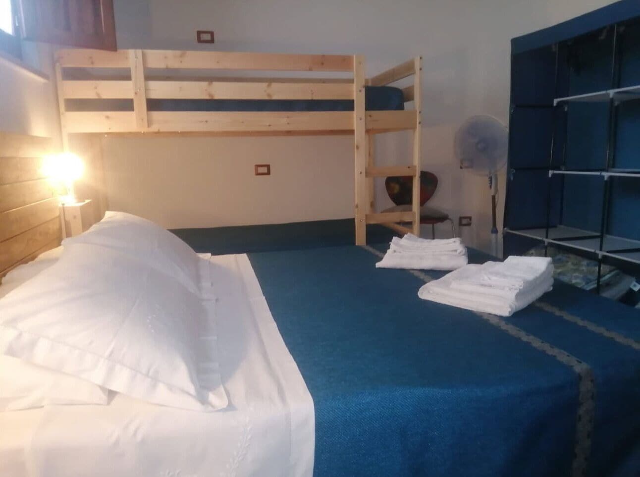 Habitación con literas, un colchón azul, almohadas blancas y un ventilador.
