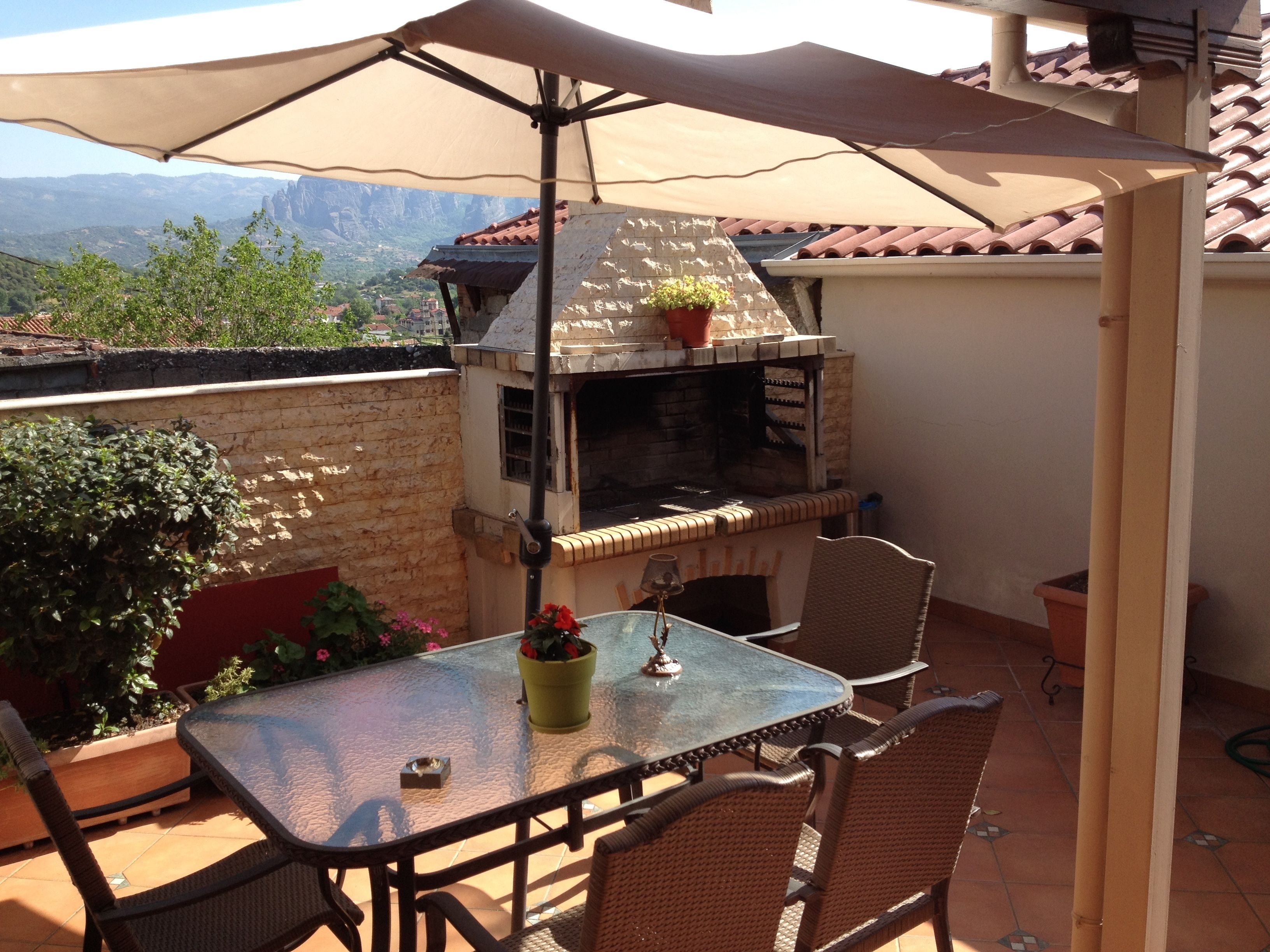 Una veranda esterna con tavolo in vetro, sedie in vimini, barbecue e vista sulle montagne.