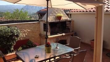 Una veranda esterna con tavolo in vetro, sedie in vimini, barbecue e vista sulle montagne.