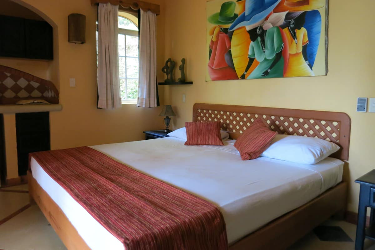 A bedroom with a large bed, a window with curtains, a painting on the wall, and a wooden headboard.