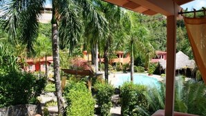 A view of a pool surrounded by palm trees and red-roofed buildings, with a stone wall and a pergola in the foreground. - Madrugada Residencial (Las Terrenas)