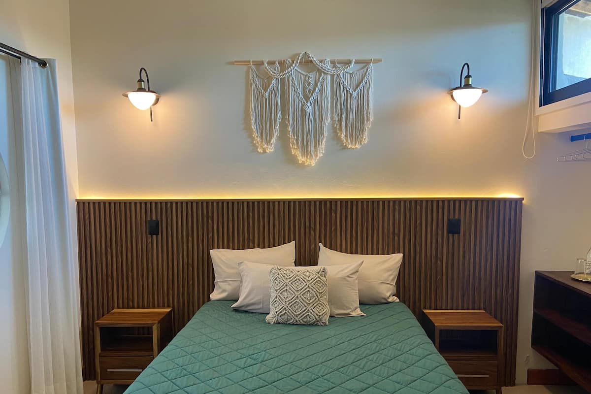 Quarto com cabeceira de madeira, colcha acolchoada verde-água, duas mesas de cabeceira e decorações de macramé fixadas na parede.