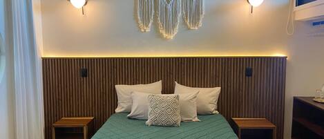 Un dormitorio con cabecera de madera, colcha acolchada color verde azulado, dos mesitas de noche y adornos de macramé en la pared.