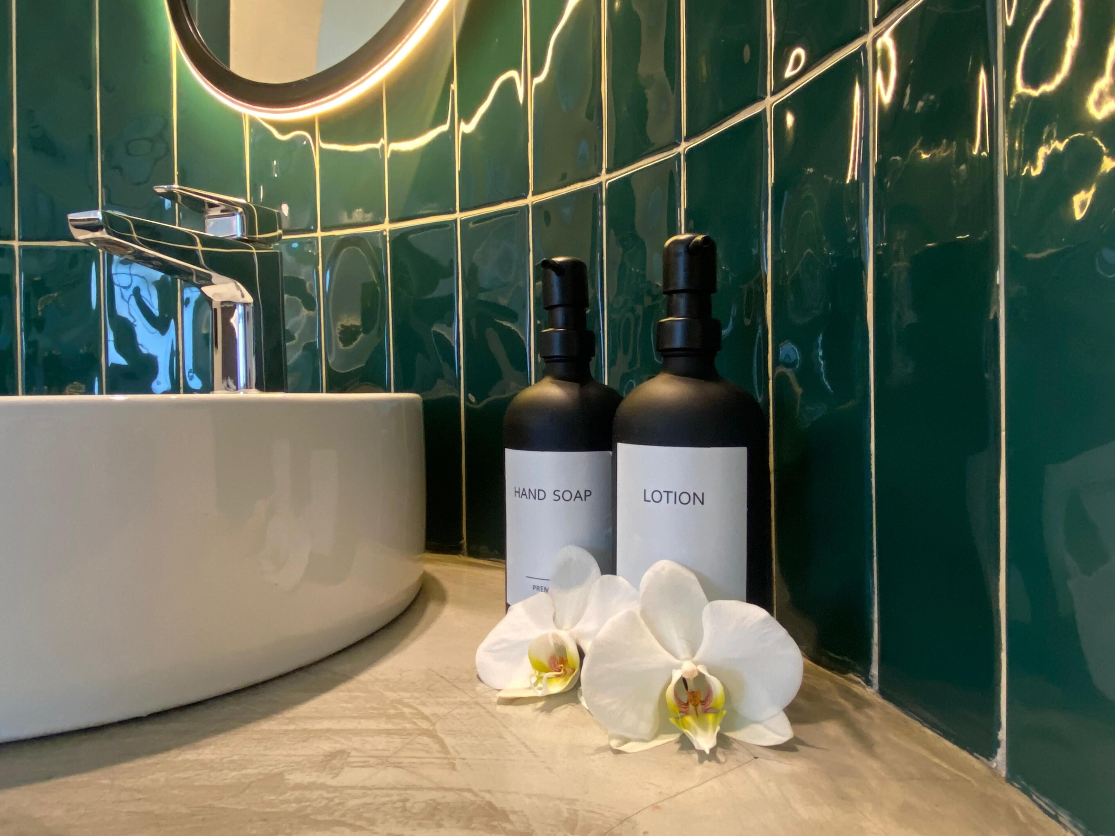 Et badeværelse med en hvid vask, to flasker mærket "HAND SOAP" og "LOTION", samt hvide orkidéer på gulvet.