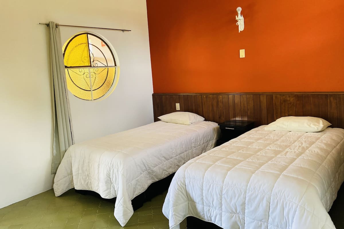 Quarto de hotel com duas camas de solteiro, cabeceira de madeira, luminária fixada na parede e janela redonda com cortina.