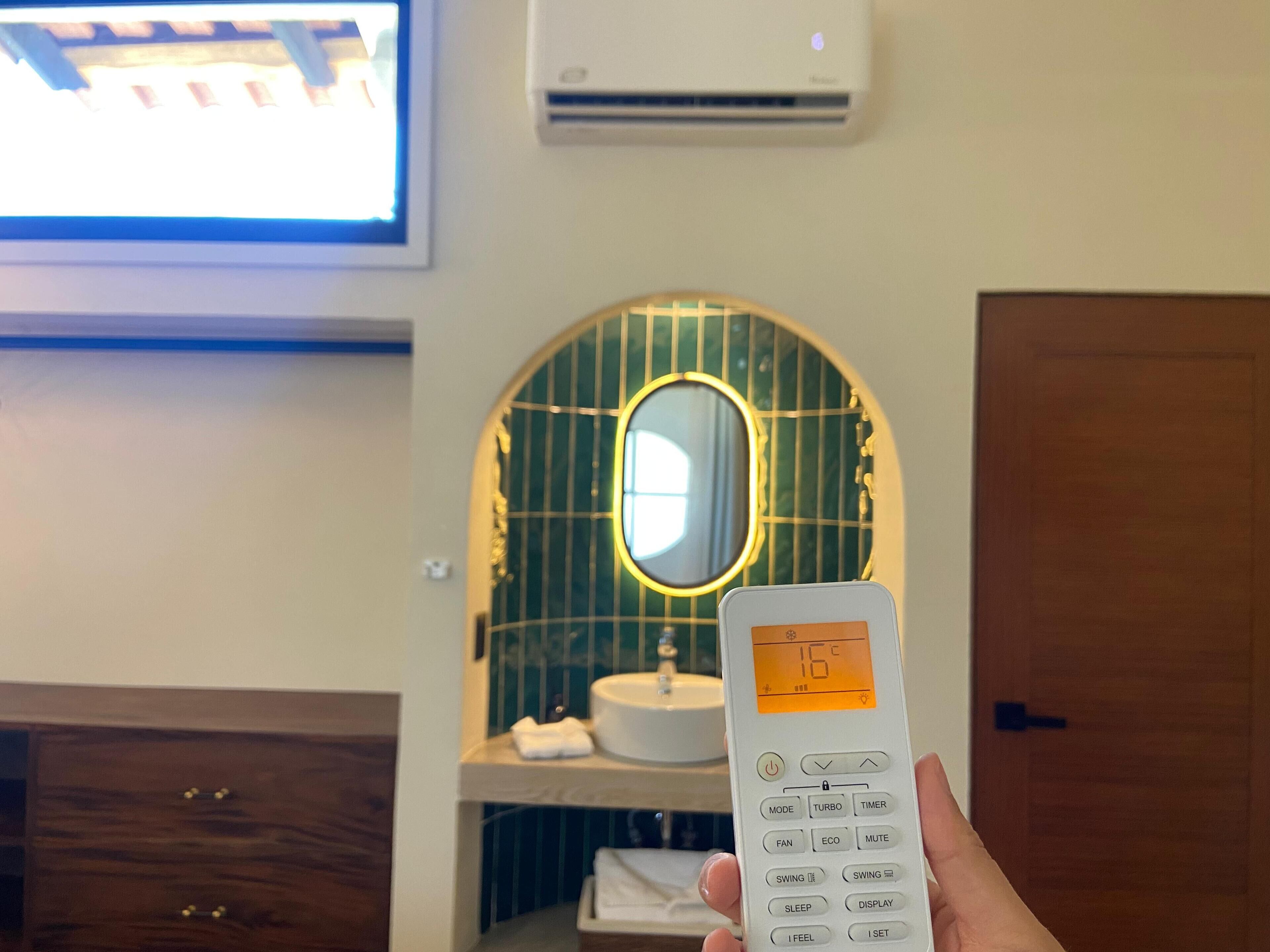 Eine Hand hält einen Thermostat mit digitaler Anzeige, darüber eine Klimaanlage, ein Holzkabinent und ein Spiegel mit dekorativem Rahmen.