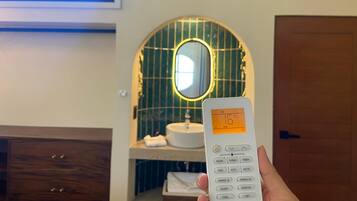 Eine Hand hält einen Thermostat mit digitaler Anzeige, darüber eine Klimaanlage, ein Holzkabinent und ein Spiegel mit dekorativem Rahmen.