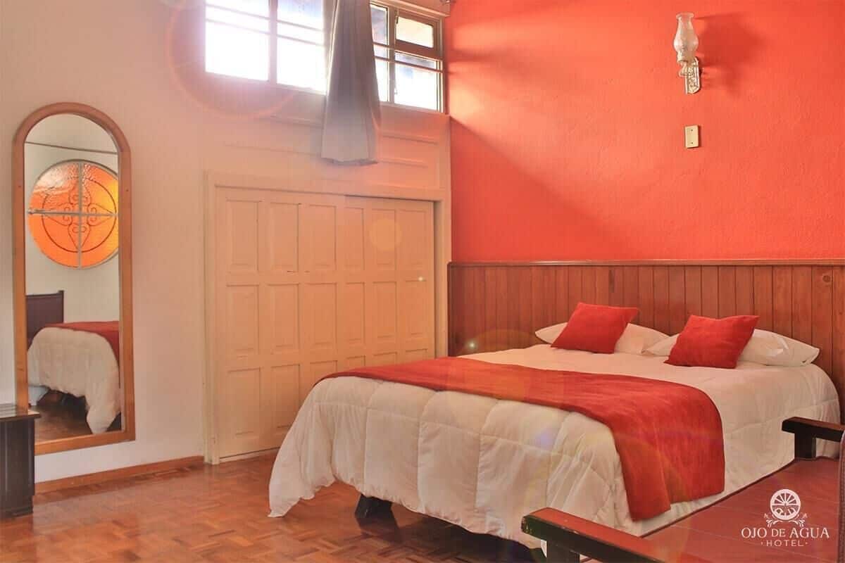 Quarto de hotel com uma cama, espelho, chão de madeira e parede vermelha.