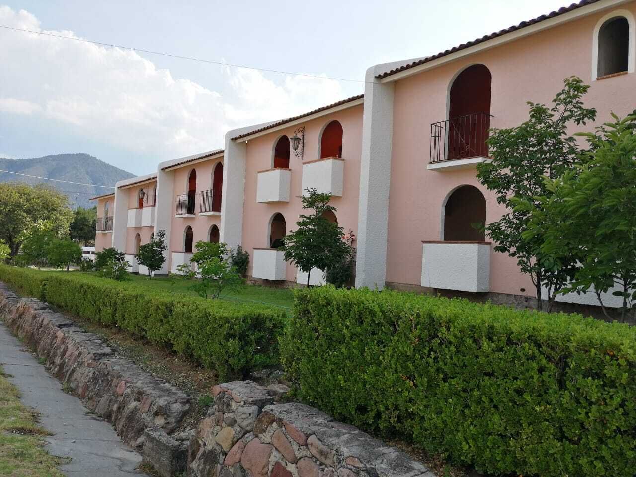 A row of pink buildings with balconies and arched windows, a stone pathway, and green hedges.