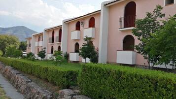 A row of pink buildings with balconies and arched windows, a stone pathway, and green hedges.