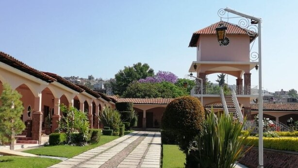A building with a covered walkway, a bell tower, and a garden with neatly trimmed hedges.