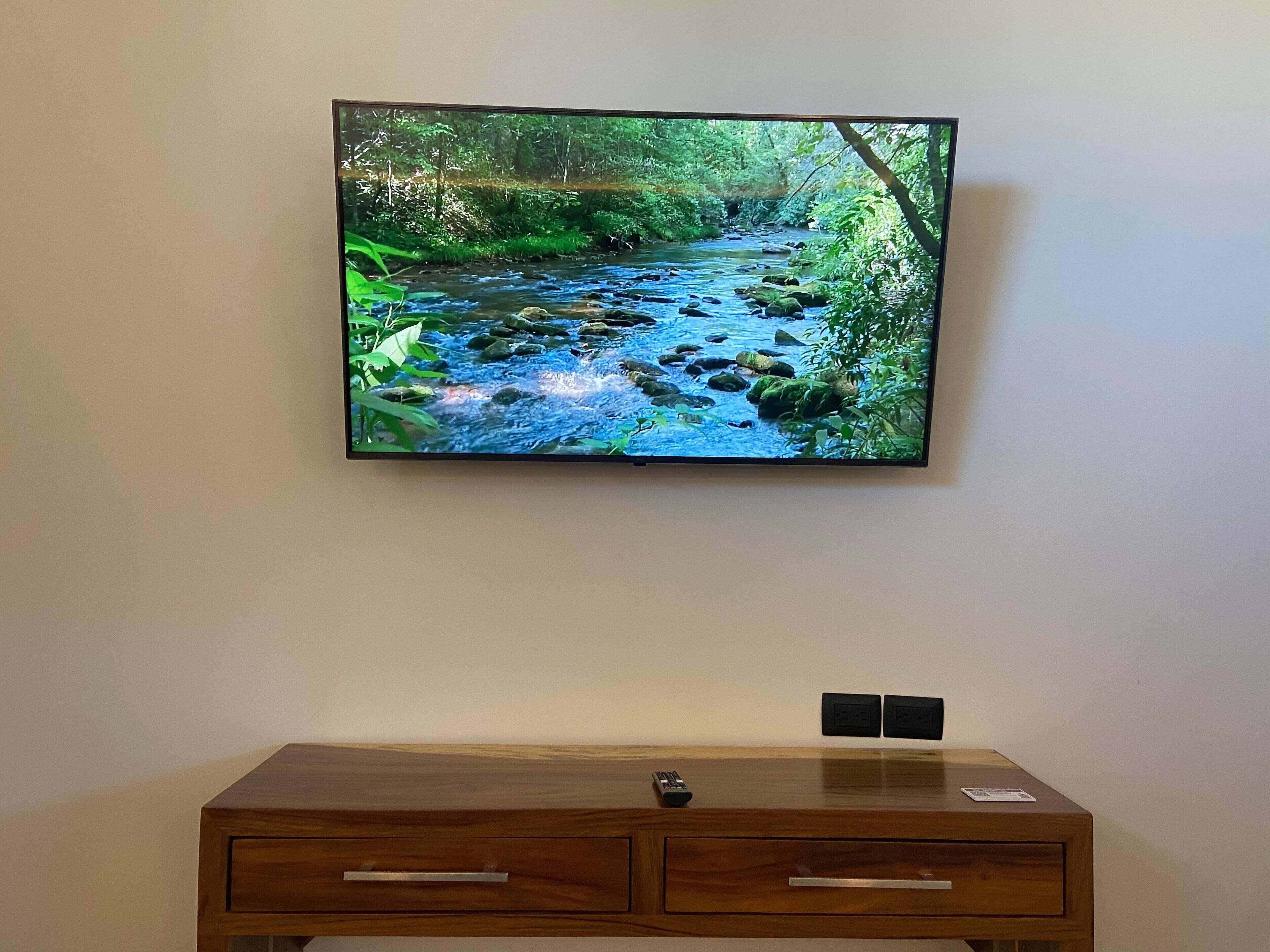 A flat-screen TV mounted on a wall displaying a natural landscape. Below the TV, there is a wooden dresser with two drawers and a remote control on top.