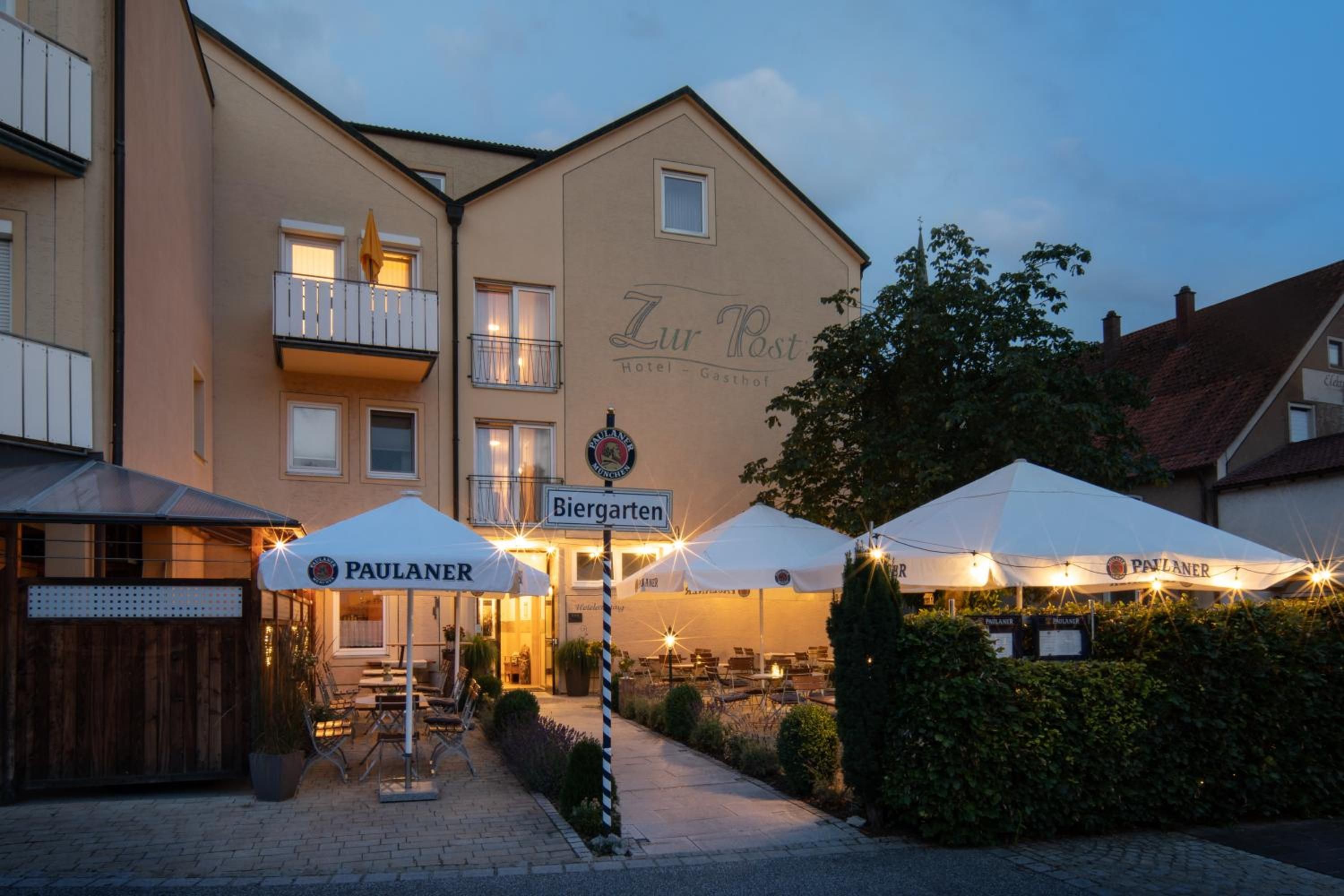 Ein Hotel namens 'Zur Post' mit Außenbestuhlung und Sonnenschirmen, einem Schild für 'Biergarten' und einem Paulaner-Bierzelteinrichtung.
