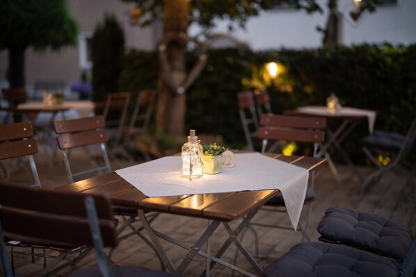 Un área de comedor al aire libre con mesas y sillas, una mesita con una vela y una planta, y un número sobre la mesa.