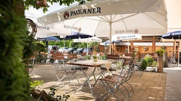 Ein Außenbereich mit Tischen und Stühlen unter Sonnenschirmen mit dem Schriftzug 'PAULANER'.