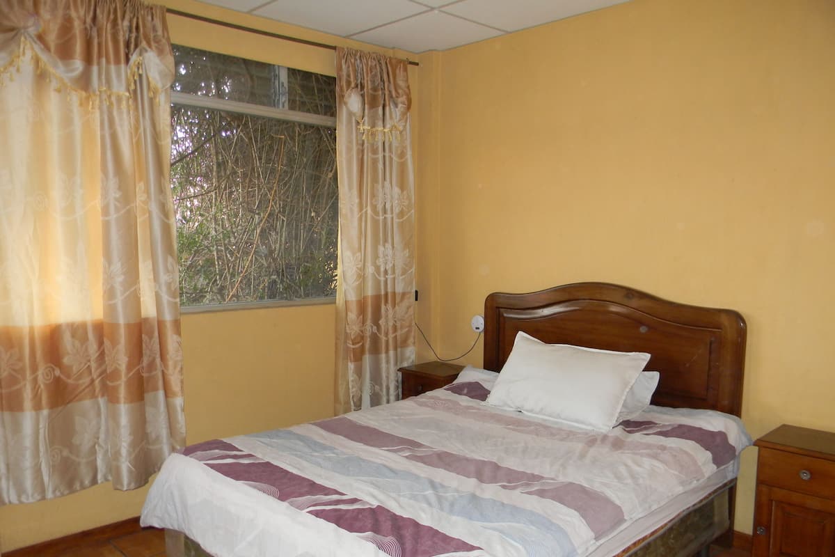 A bedroom with a wooden bed, a nightstand, a window with curtains, and a tiled floor.