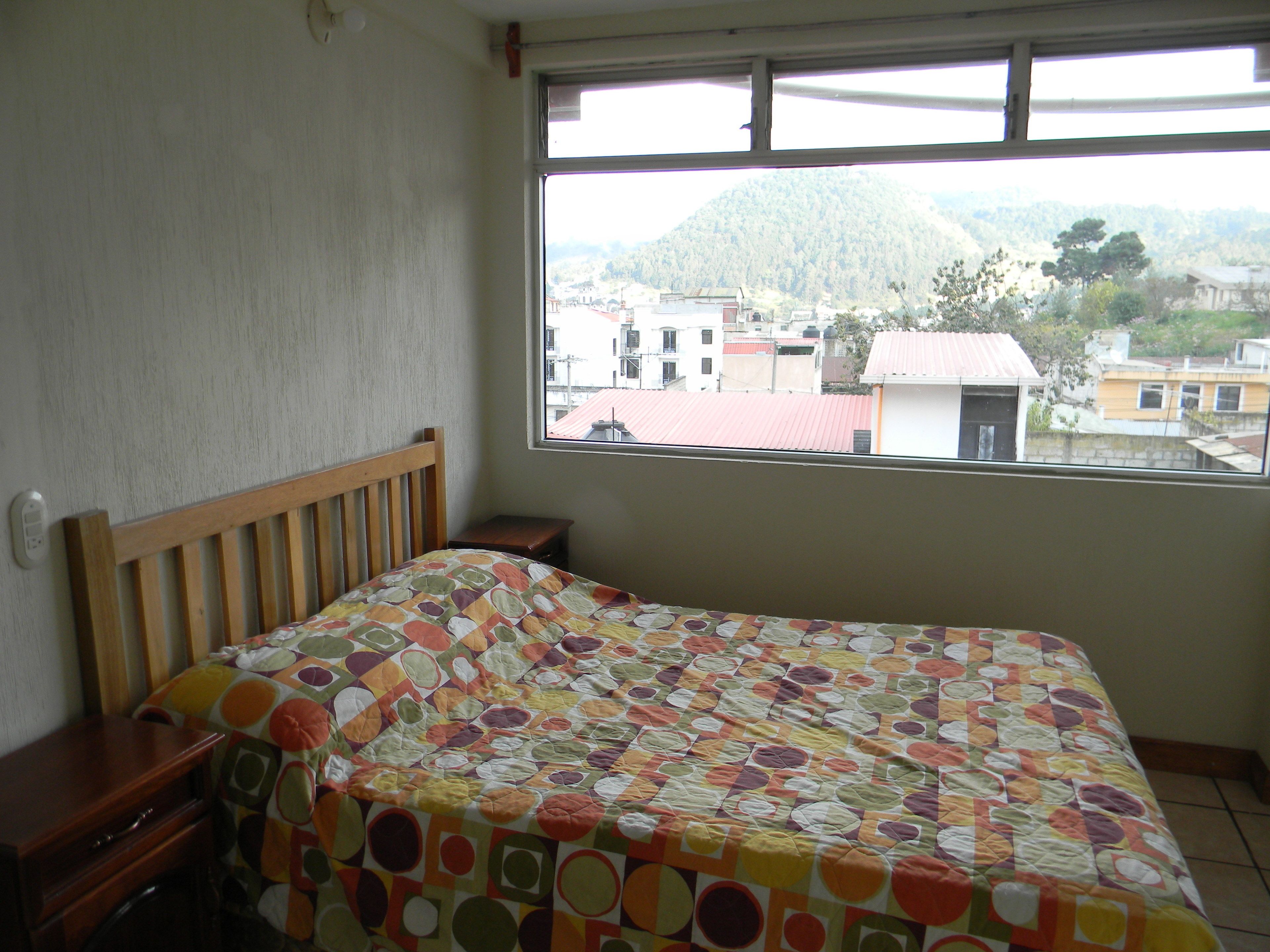 A bedroom with a wooden bed, a small bedside table, and a window with a view of buildings and hills.