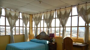 Une chambre avec un lit, un bureau et une chaise. Les fenêtres sont dotées de rideaux et la pièce est recouverte de carrelage.