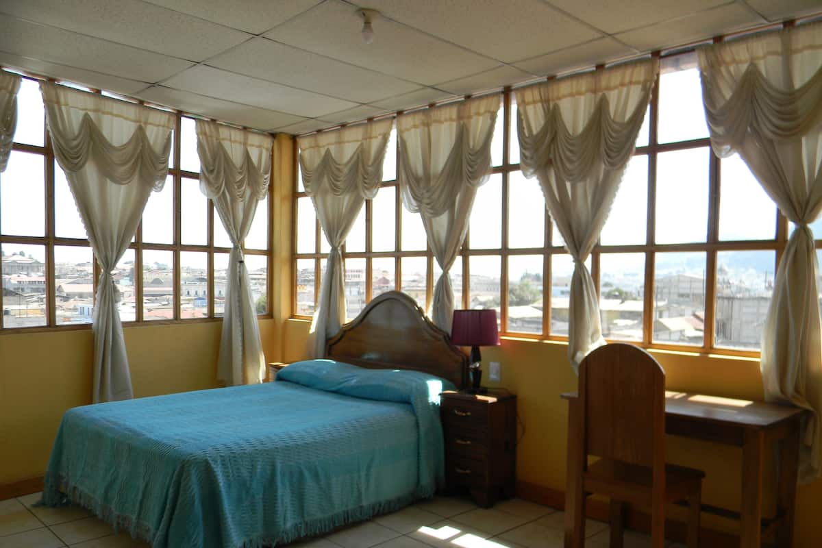 A bedroom with a bed, a desk, and a chair. There are curtains on the windows, and the room has a tiled floor.