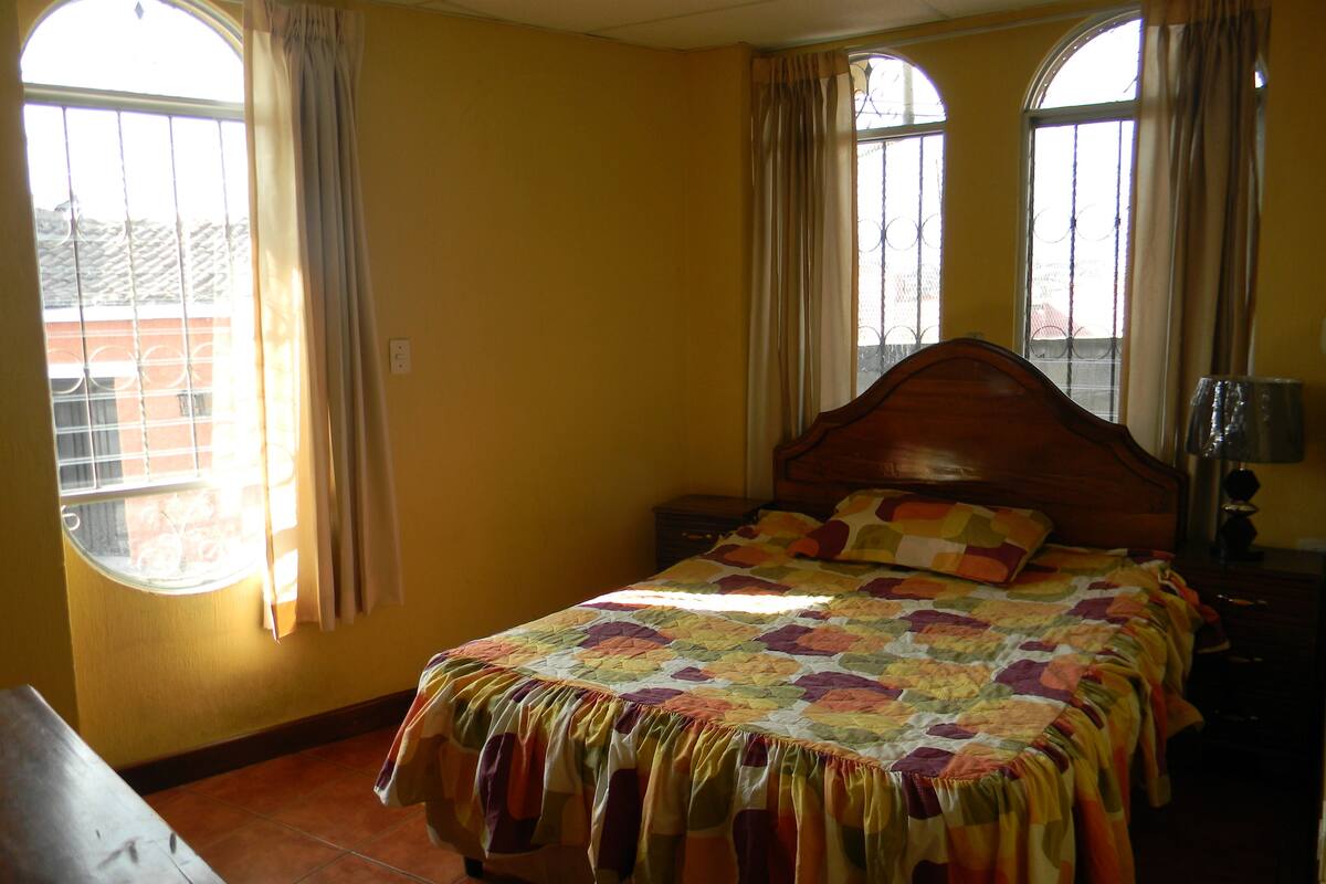 A bedroom with a wooden bed, a nightstand, a lamp, and a window with curtains.