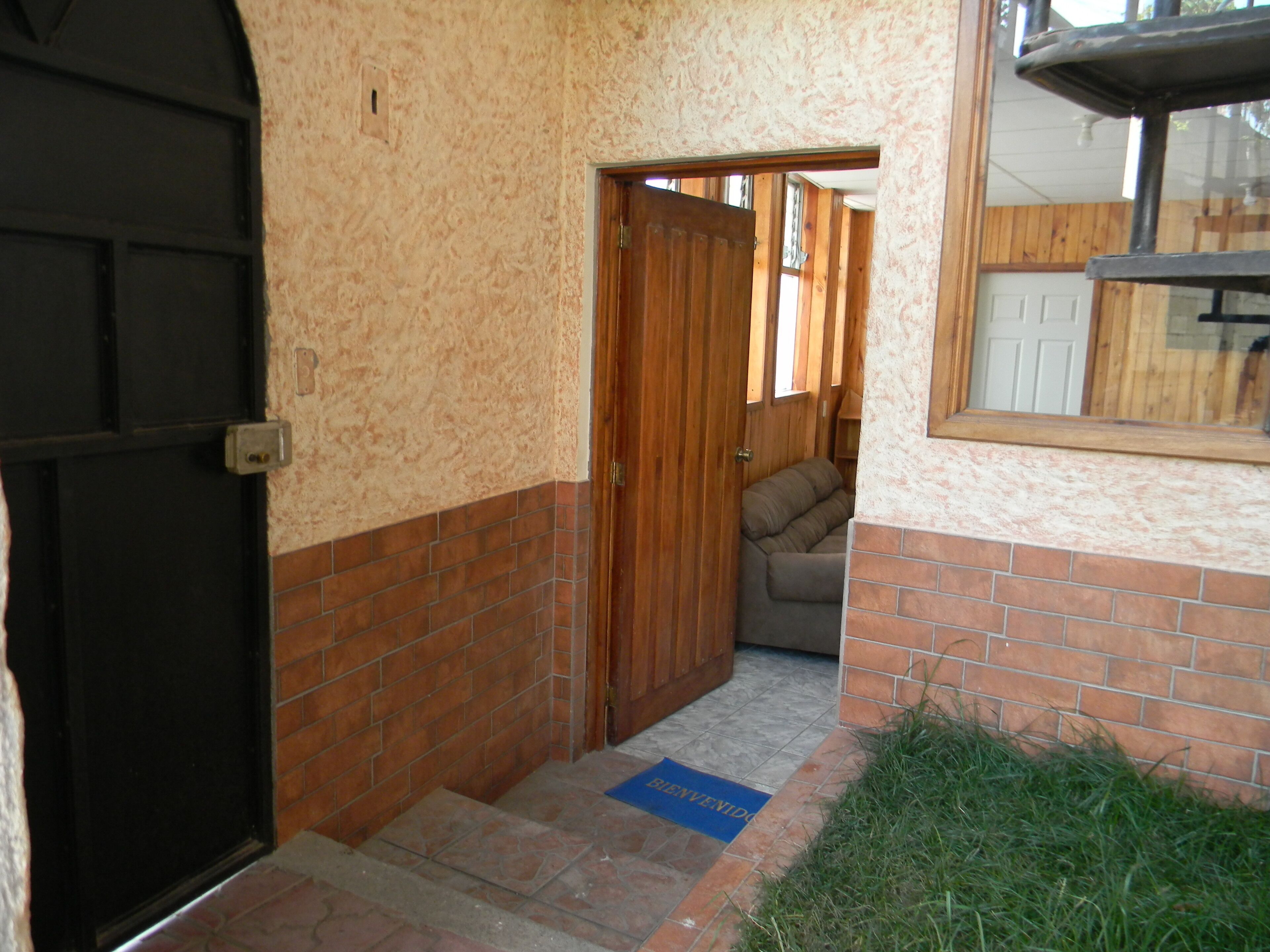Un pasillo estrecho con una puerta negra, una puerta de madera que da a una habitación y un tapete azul con la palabra "BIENVENIDOS".
