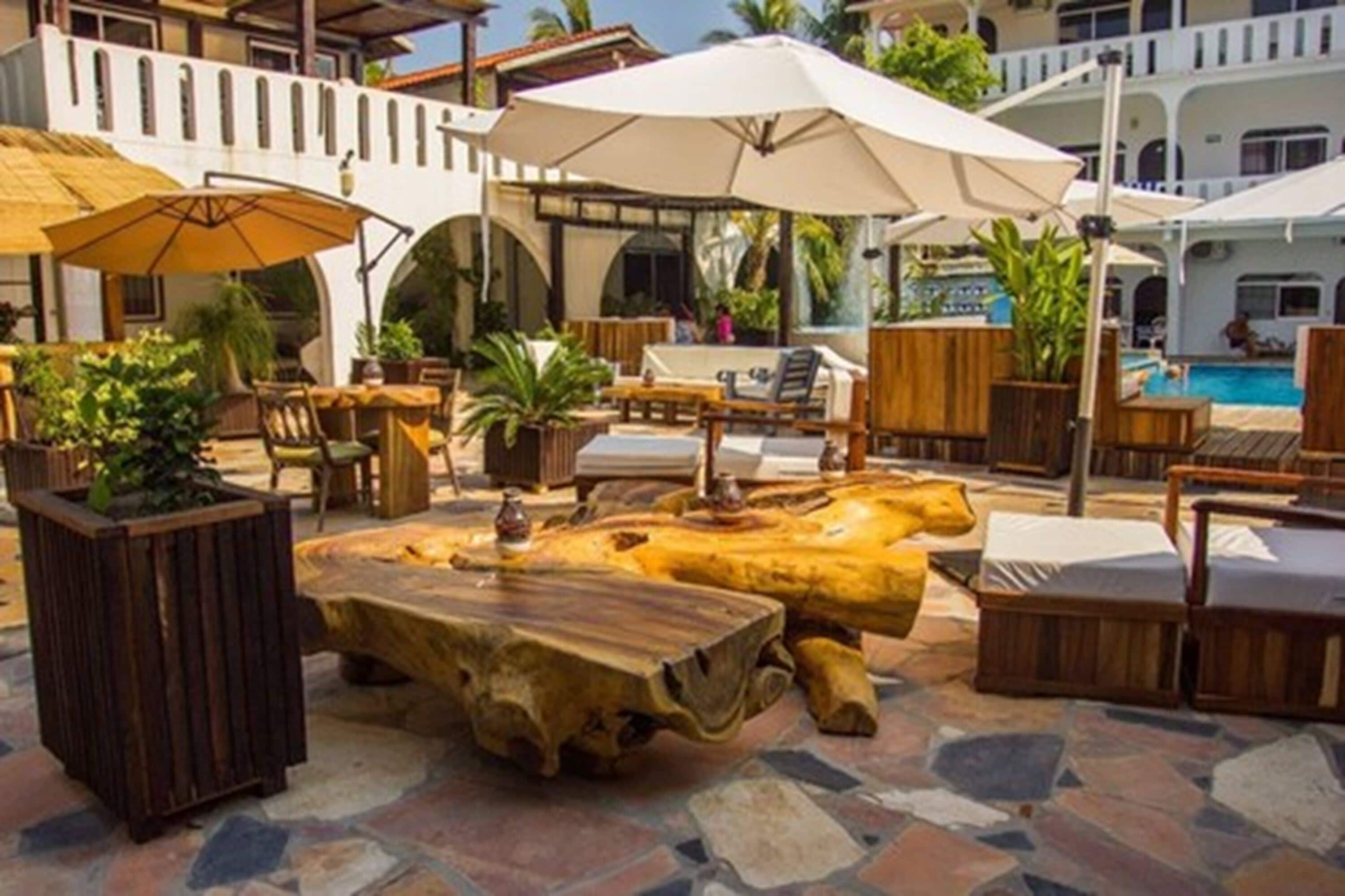 An outdoor lounge area with wooden furniture, umbrellas, and a pool in the background.