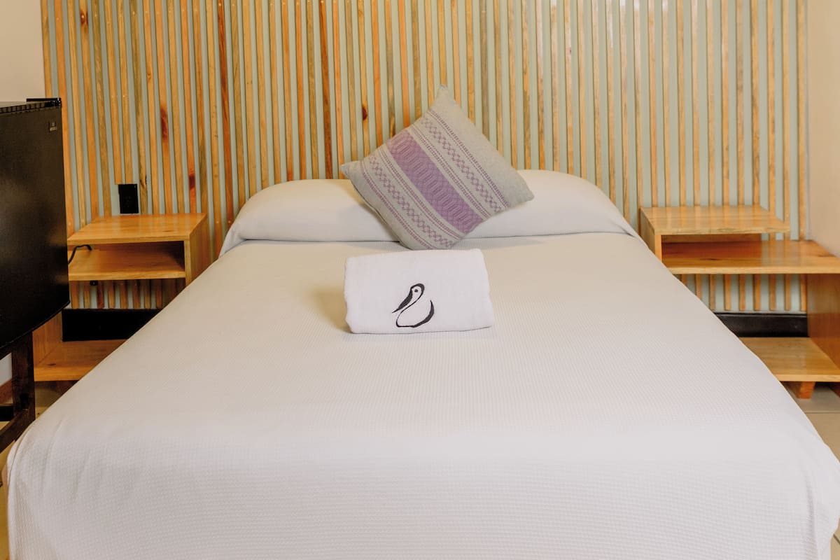 Una cama bien hecha con una almohada y una toalla, mesitas de noche de madera y un televisor a la izquierda.