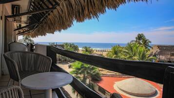 Varanda com cadeiras de vime e uma mesa pequena, com vista para uma praia com palmeiras e um edifício de telhado vermelho.