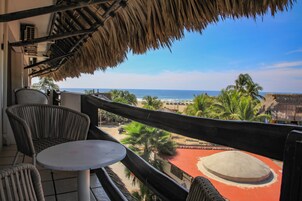 Un balcón con sillas de mimbre y una pequeña mesa, con vistas a una playa con palmeras y un edificio de techo rojo.