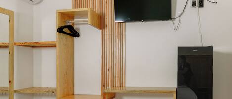A room with a TV mounted on the wall, a wooden shelf unit, and a refrigerator.