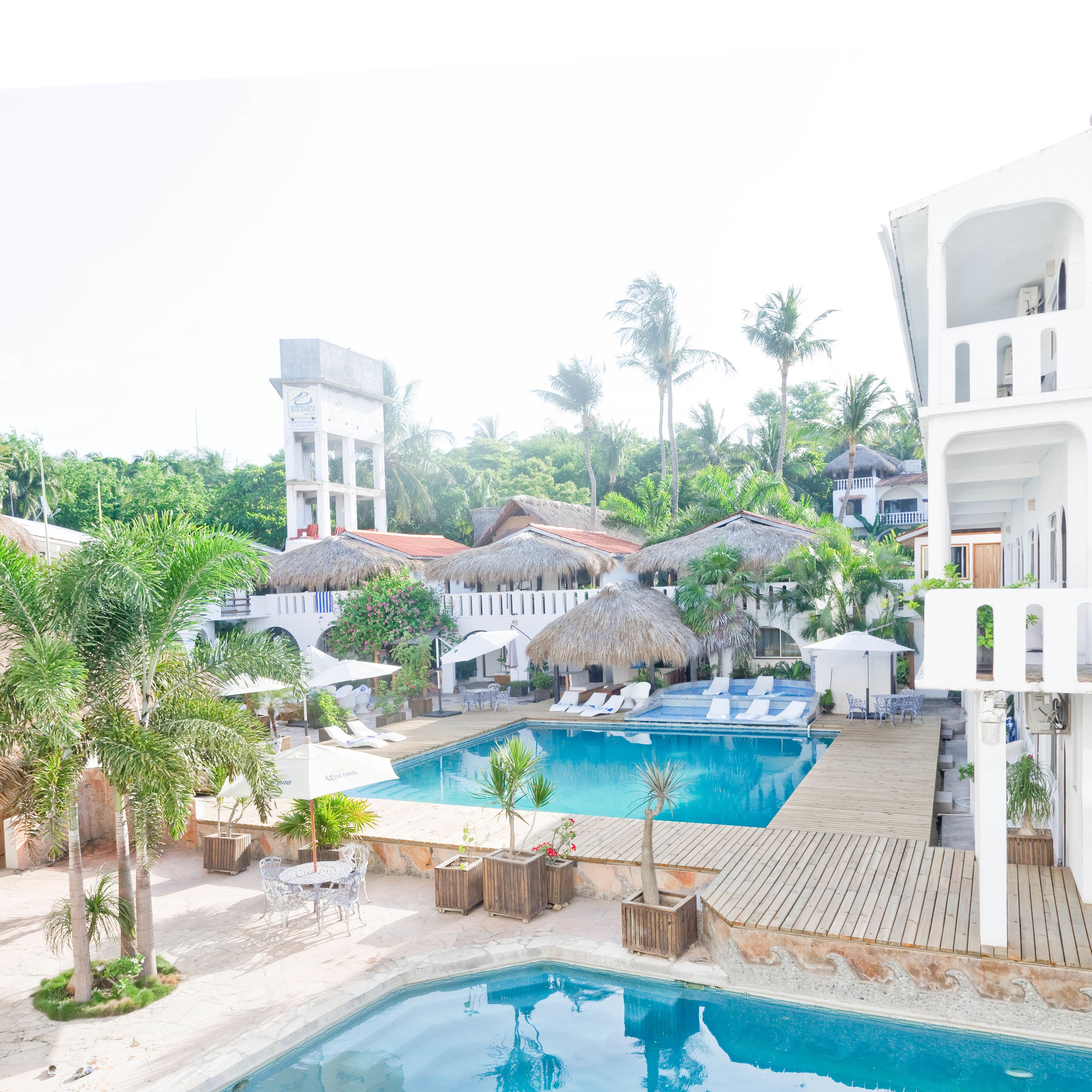Un resort con piscina central, edificaciones de techo de paja y un edificio blanco con balcones.