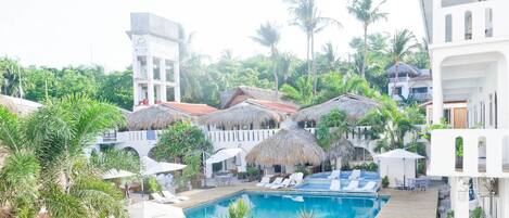 Un resort con piscina central, edificaciones de techo de paja y un edificio blanco con balcones.