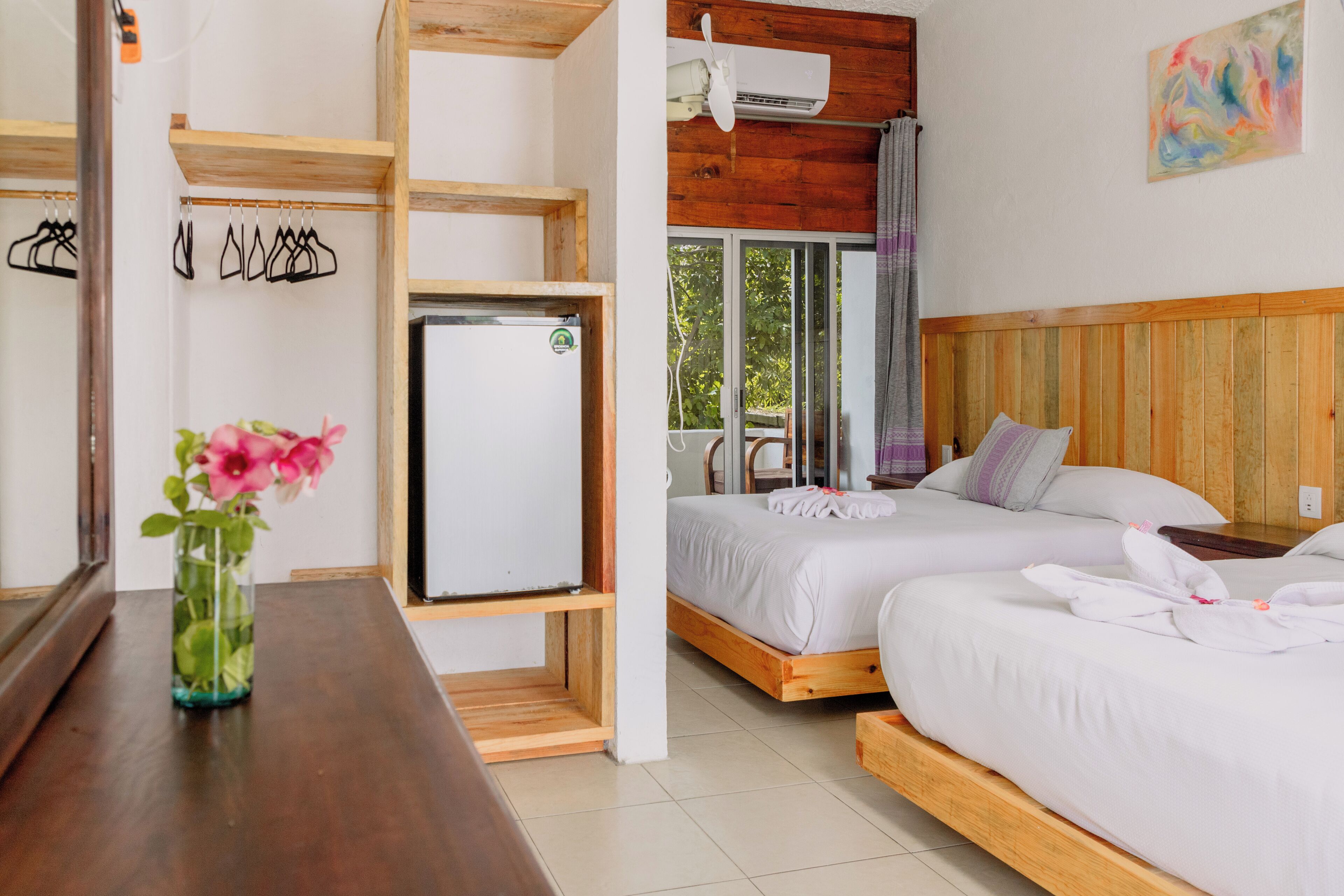 A hotel room with two beds, a wooden headboard, a small table with a vase of flowers, a wall-mounted air conditioner, and a window with a view of greenery.