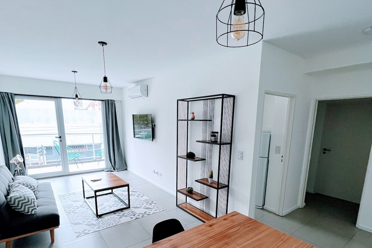 A modern living room with a wooden dining table, black chairs, a black sofa, a patterned rug, and a black metal shelving unit.