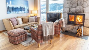 Una sala de estar acogedora con chimenea de piedra, un sofá, sillones a cuadros y un ventanal con vistas nevadas.