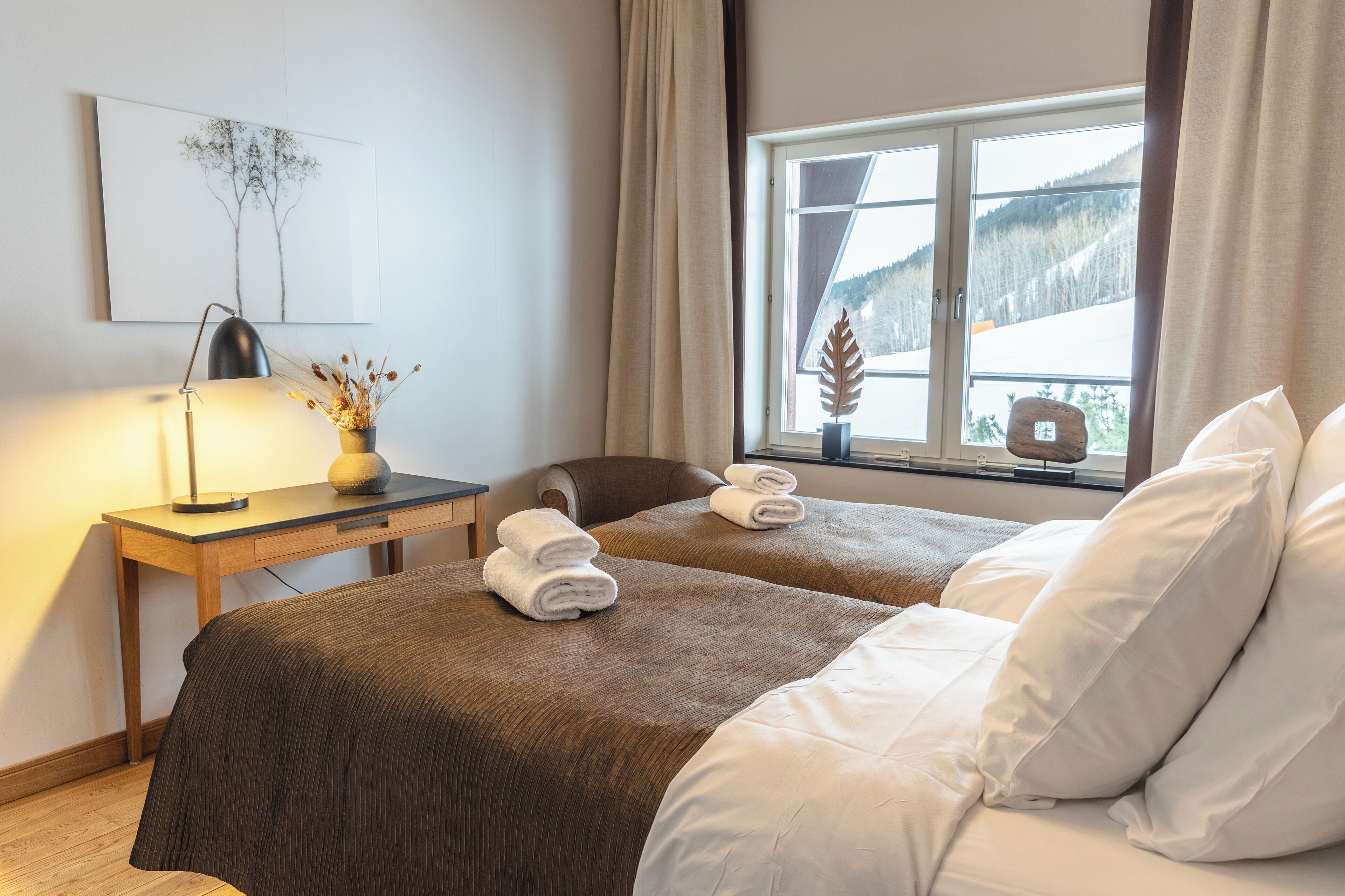 A hotel room with two beds, a desk, a lamp, and a window with a view of a snowy landscape.