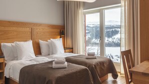 Habitación de hotel con cabecera de madera, dos camas con ropa de cama blanca y marrón, una mesa de madera y vistas a un paisaje nevado a través de un gran ventanal.