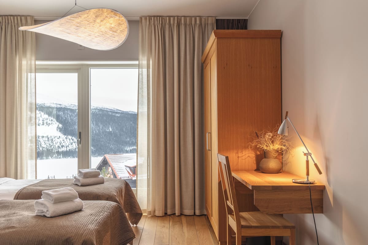 Une chambre d’hôtel avec deux lits, un bureau avec une lampe et une vue sur des montagnes enneigées.